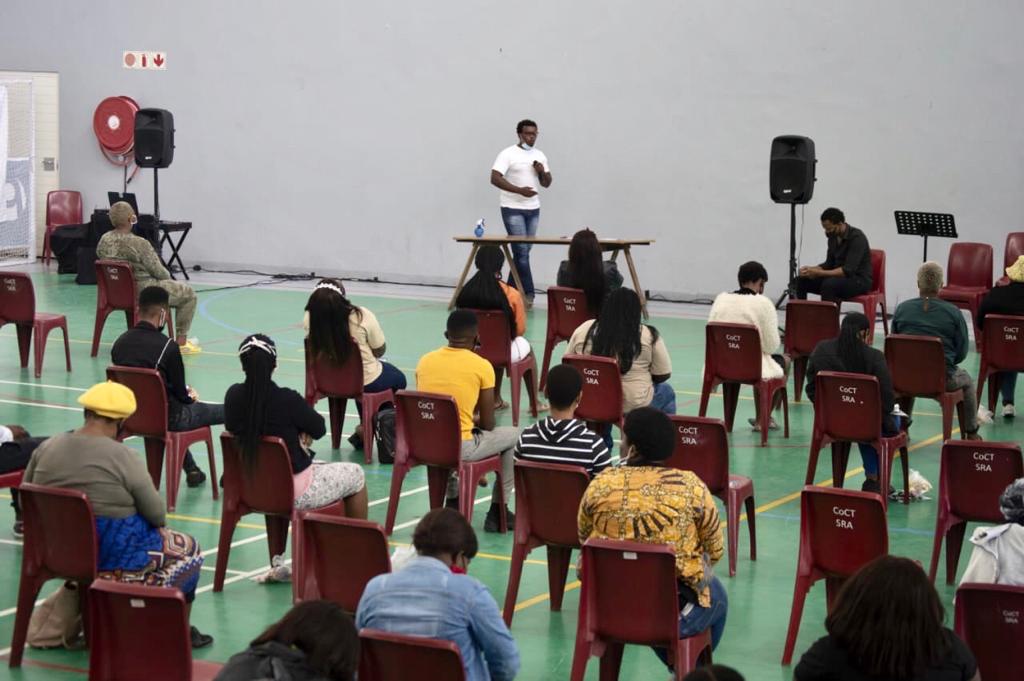 A group of socially-distanced people listen to a speaker at the front of the room.