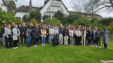 A groups of people standing in a garden on a green lawn in front of a white building 