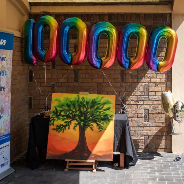 Multi-coloured balloons in the shape of the number "100 000" float against a brick wall. Underneath is a painting of a tree. To the left is a banner showing the C-SHARP acronym and its meaning.