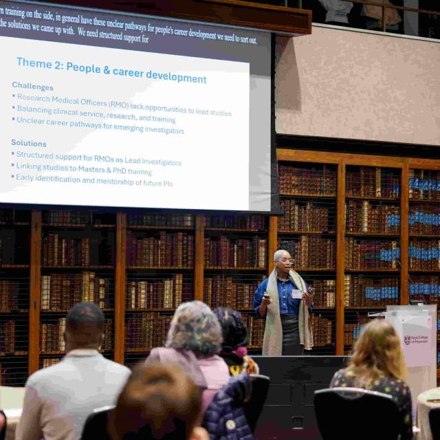 Prof. Molebogeng Rangaka speaks in front of an audience in a library. On the screen behind her, a slide displays the text "Theme 2: People and career development"