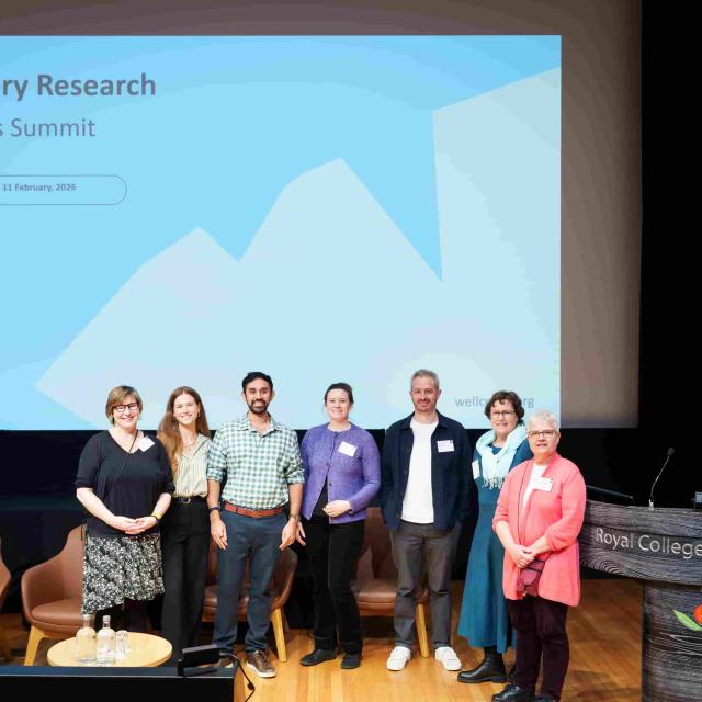 The organising and facilitation team of seven people stand at the front of an auditorium in front of a screen displaying the words "Discovery Research Platforms Summit"