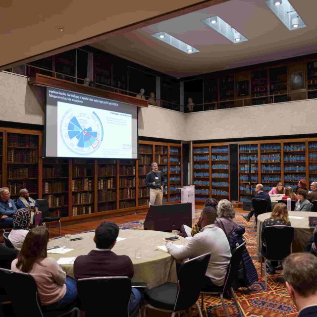 Prof. Andrew Boulle presenting to an audience in a library; behind him a slide displays a radial graph