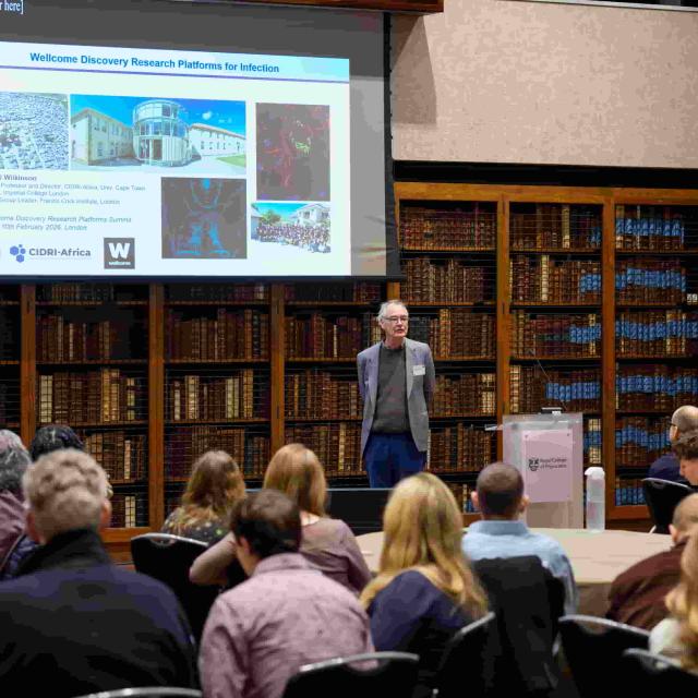 Prof. Robert J Wilkinson speaking to an audience in a library. Behind him is projected a slide titled "Wellcome Discovery Research Platform for Infection"